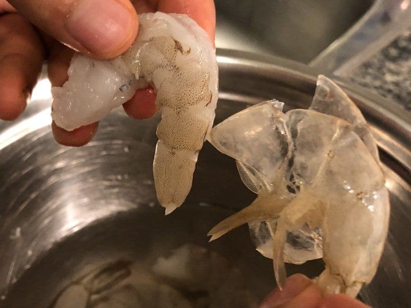 Hand holding peeled shrimp with tail removed, showing the transparent flesh and red digestive tract vein visible down the back.
