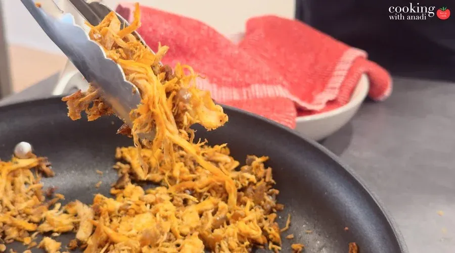 Tongs lifting seasoned shredded chicken mixture from a black skillet, golden and caramelized with spices.