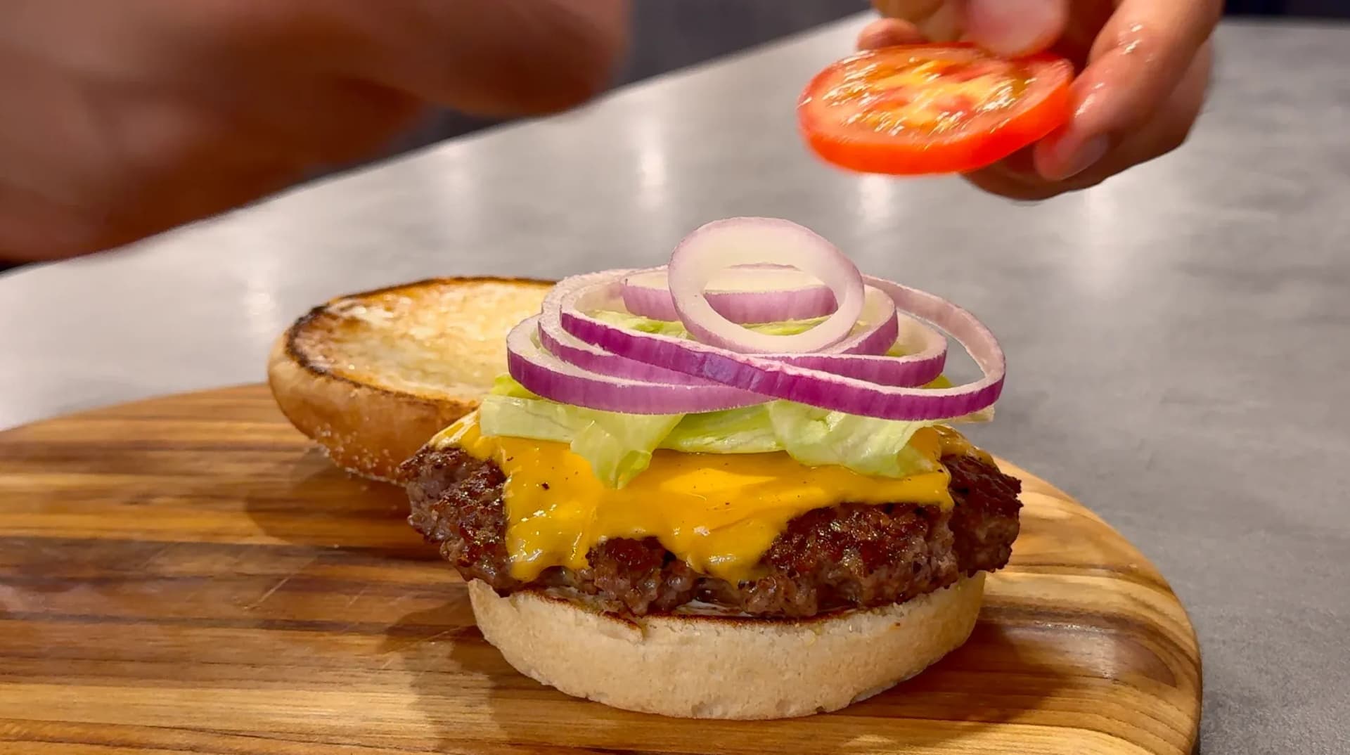 Classic cheeseburger on toasted bun with melted cheddar, lettuce, pickles, and purple onion rings, with tomato slice being added.