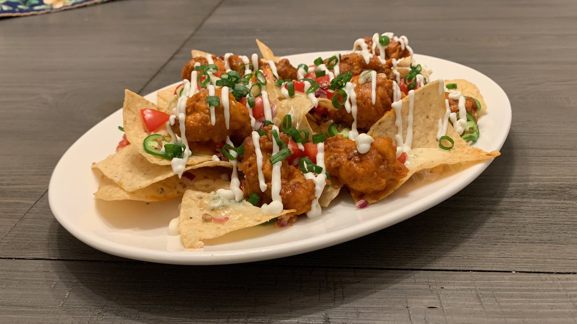 Buffalo popcorn chicken nachos topped with white drizzle sauce, diced tomatoes, and sliced jalapeños on a white plate.
