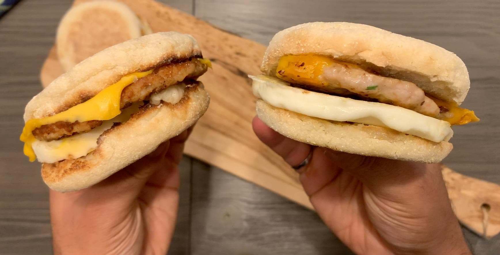 Two homemade breakfast sandwiches held in hands, featuring sausage patties, melted cheese, and toasted English muffins.