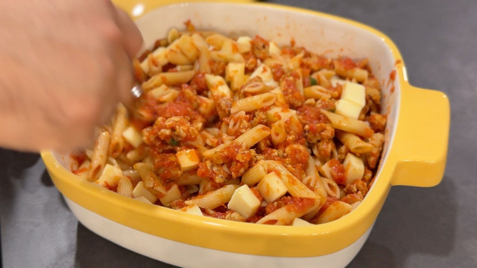 Cheesy baked penne with spicy Italian sausage in a yellow casserole dish before baking, with melted mozzarella cubes visible.
