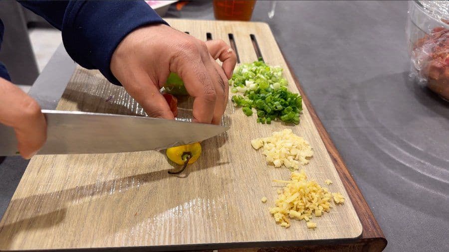 Hand chopping fresh green onions on a wooden cutting board with minced garlic and ginger nearby.