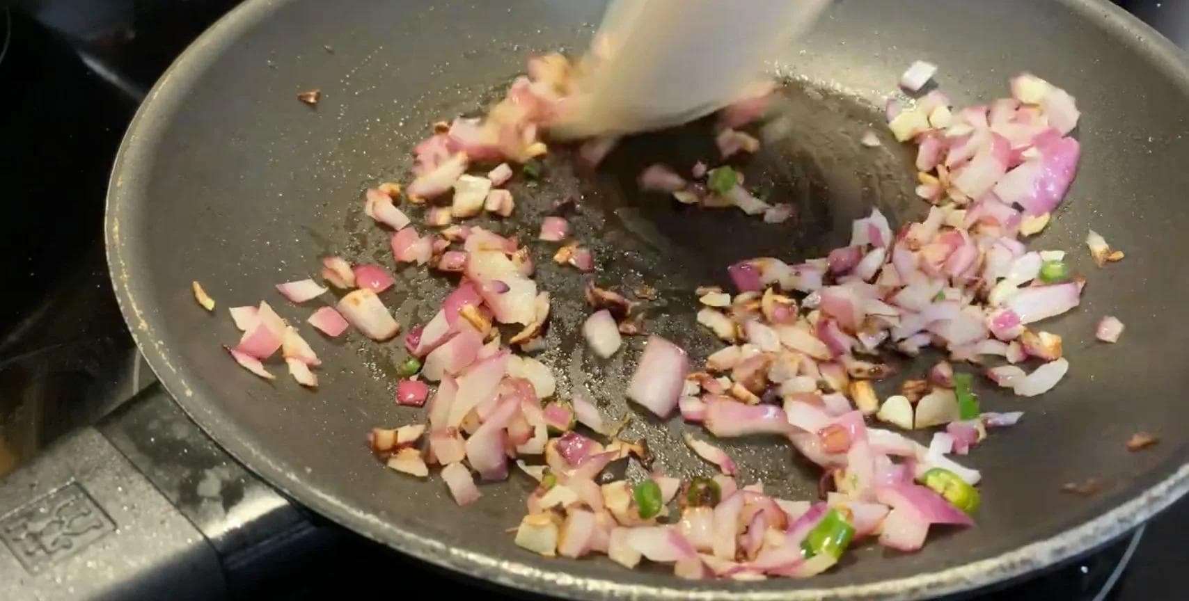Chopped onions, garlic, and chillies sautéing in a pan with a spatula mixing the ingredients.