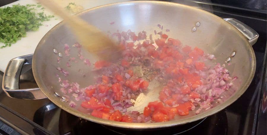 Diced tomatoes and onions cooking in a wok with spices, being stirred with a wooden spoon during tomato masala preparation.