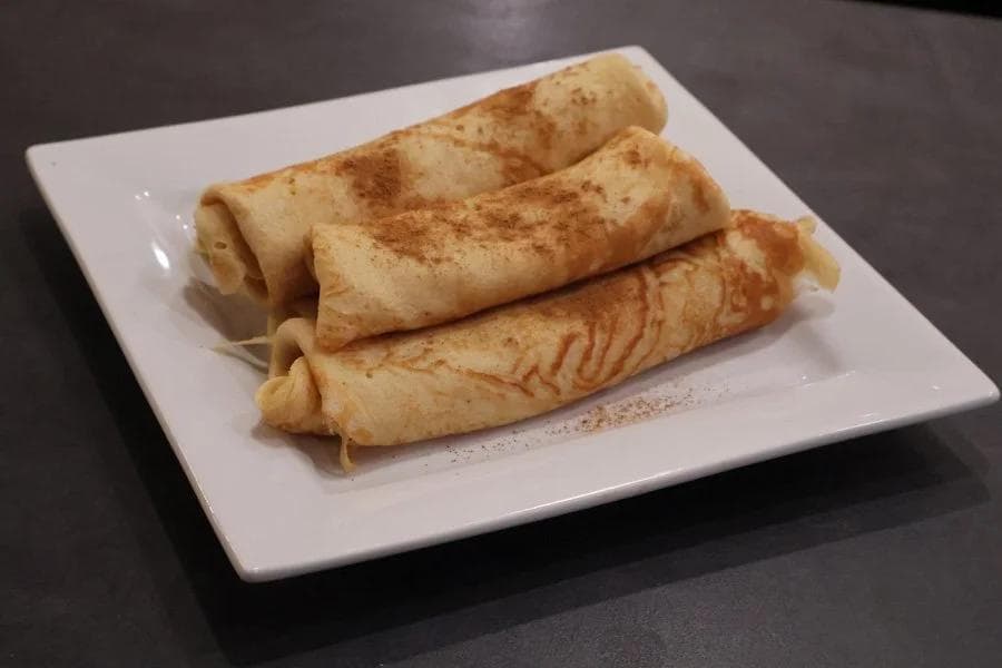 Three rolled pumpkin crepes dusted with cinnamon on a white square plate.