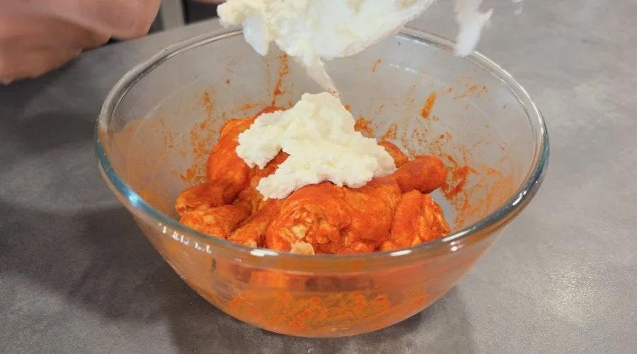 White yogurt being added to orange tandoori marinade in a glass bowl for mixing.