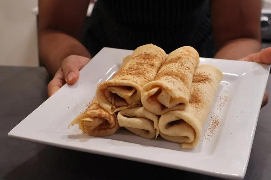 Rolled pumpkin crepes dusted with cinnamon on a white plate, held by hands.