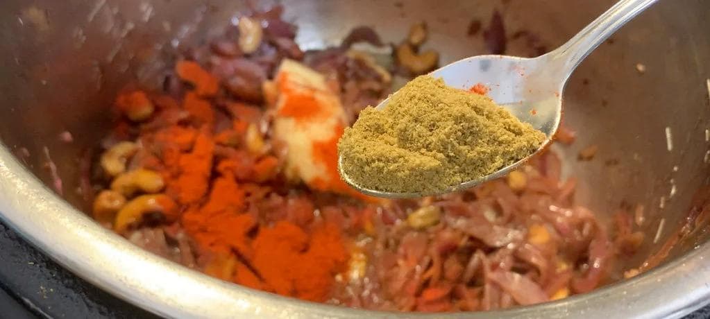 Spoon of ground coriander powder being added to cooked onions and spices in a pot for Chicken Changezi.