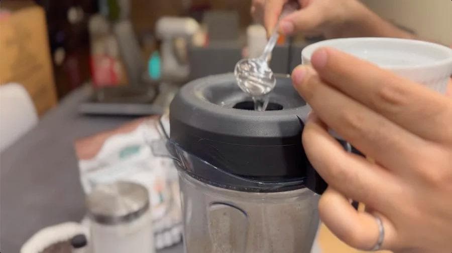 Pouring liquid coconut oil into a food processor containing ground hazelnuts while making homemade vegan Nutella.