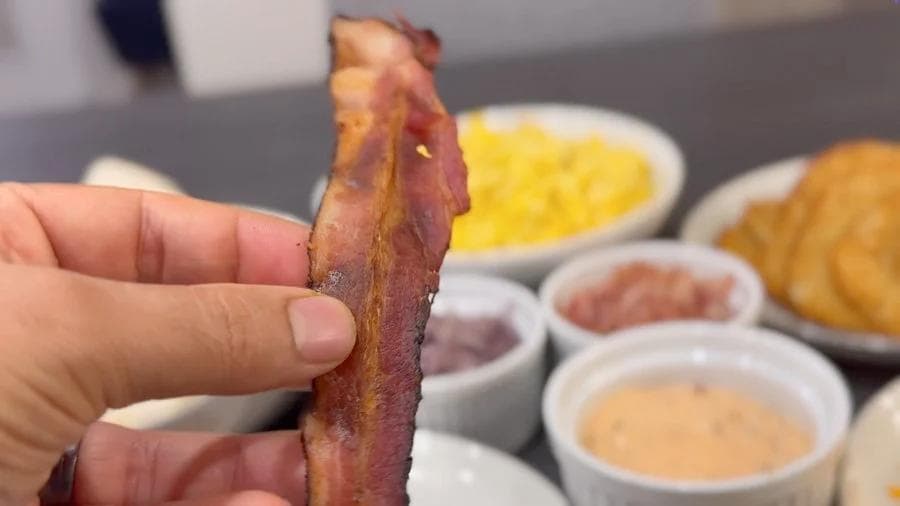 Hand holding a strip of crispy bacon with golden brown edges over bowls of breakfast burrito ingredients.