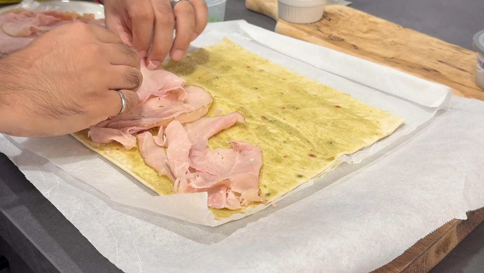 Hands layering sliced ham over a yellow herb-flecked pastry sheet on parchment paper during pinwheel assembly.