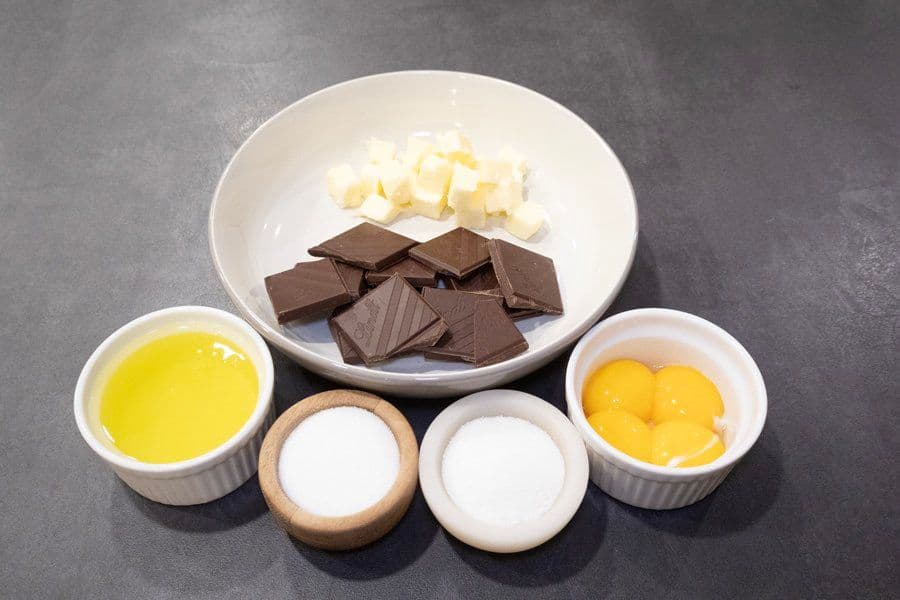 Chocolate mousse ingredients arranged on a gray surface: butter, dark chocolate pieces, oil, eggs, egg yolks, and sugar in white bowls.