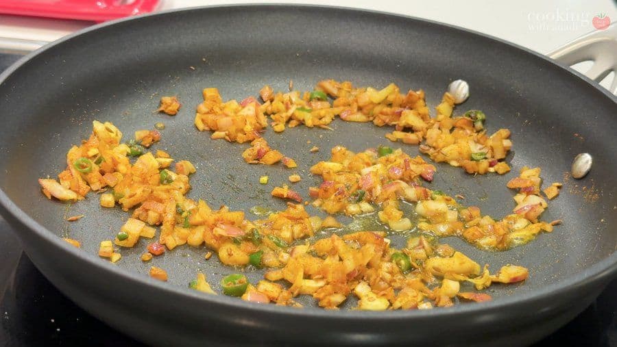Spiced potato and onion mixture cooking in a dark pan with turmeric, coriander, and green herbs visible.