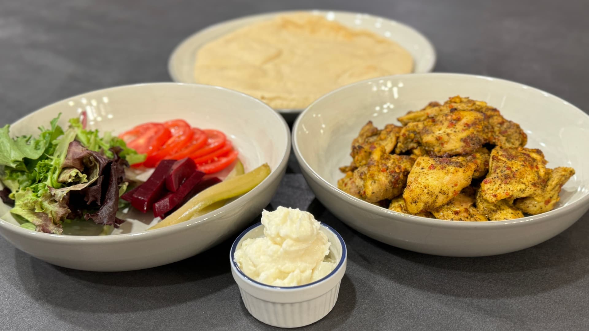 Chicken shawarma plated with fresh vegetables, hummus, spiced chicken pieces, and pita bread on a dark surface.
