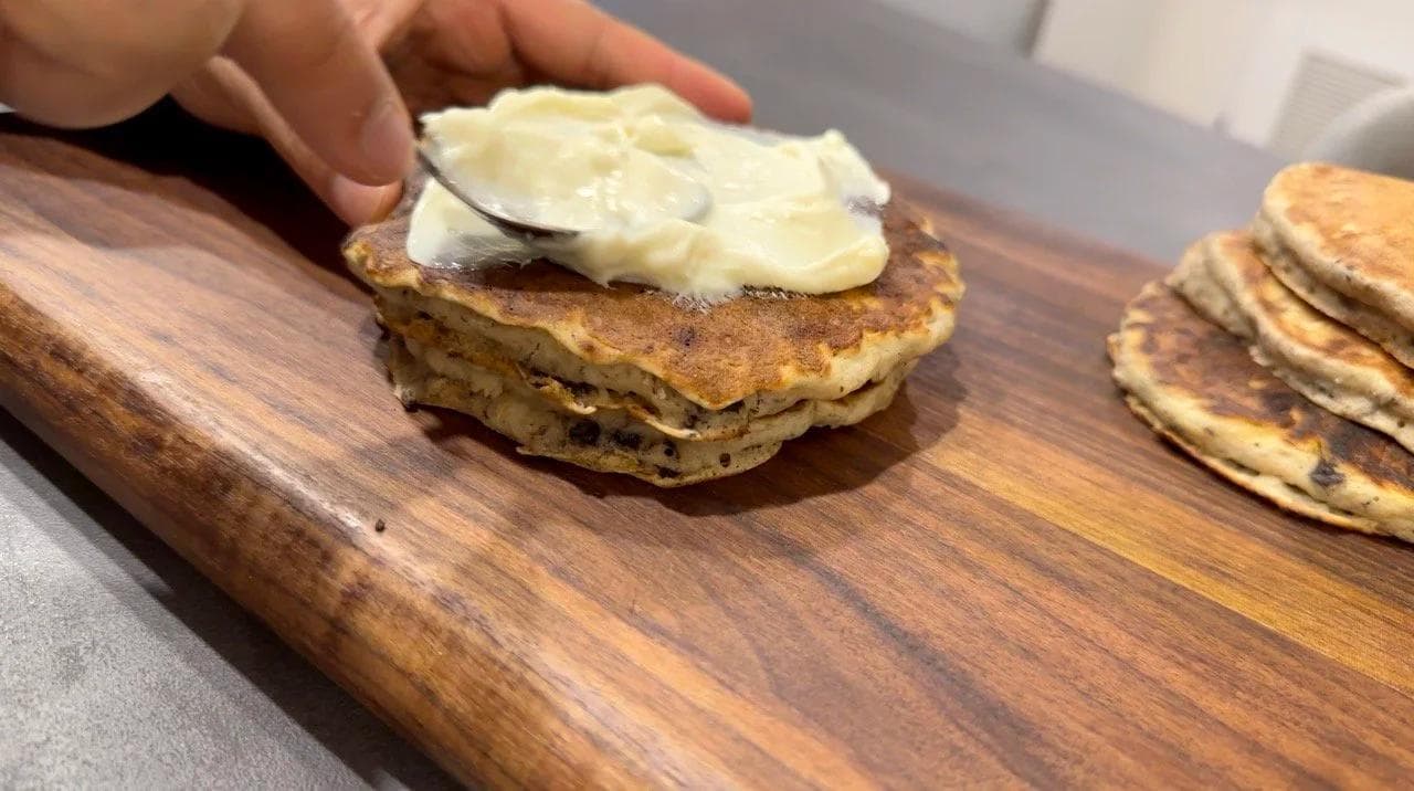 Hand spreading cream cheese glaze on a stack of gluten-free pancakes on a wooden board.