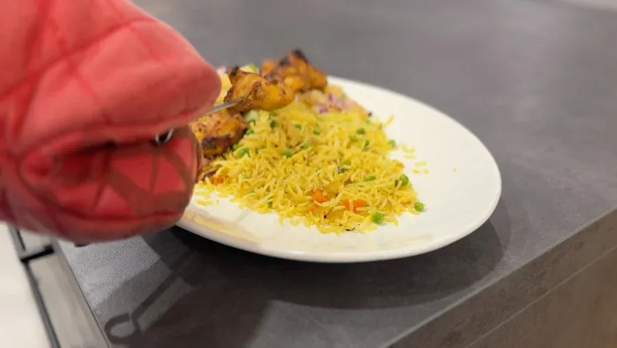 Singapore Chicken Satay served on white plate with yellow rice, cucumber, and pineapple salsa, hand holding red napkin.