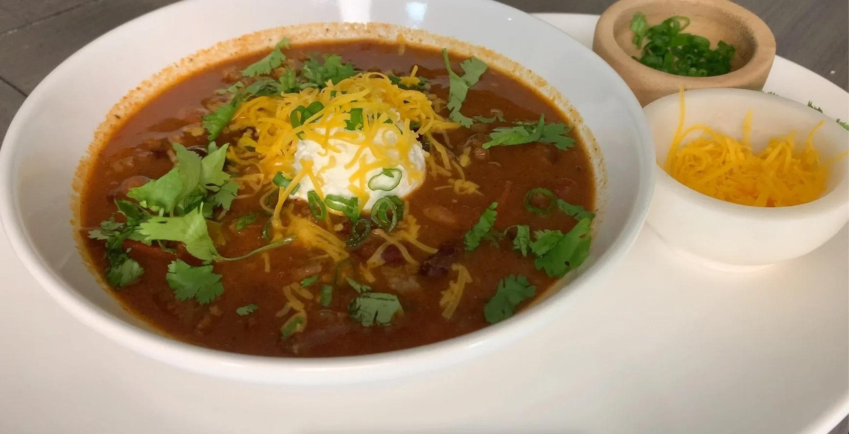 Bowl of beef chili topped with shredded cheese, sour cream, green onions, and cilantro, with side bowls of cheese and garnishes.