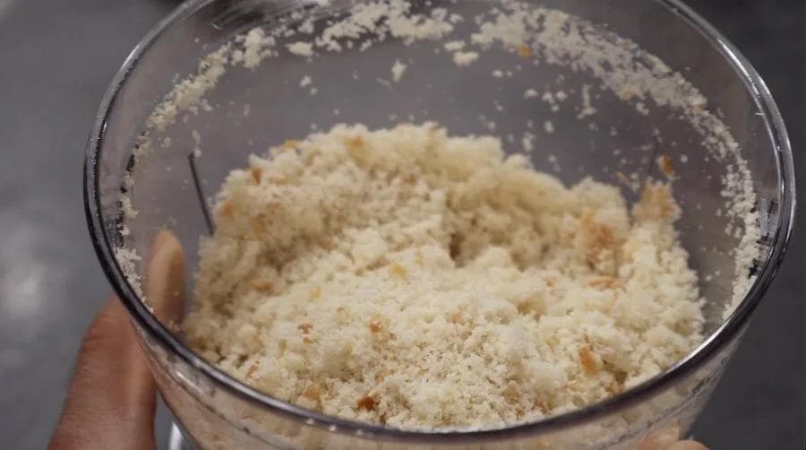 Breadcrumb coating mixture in a food processor bowl with crumbly texture for chicken nuggets.
