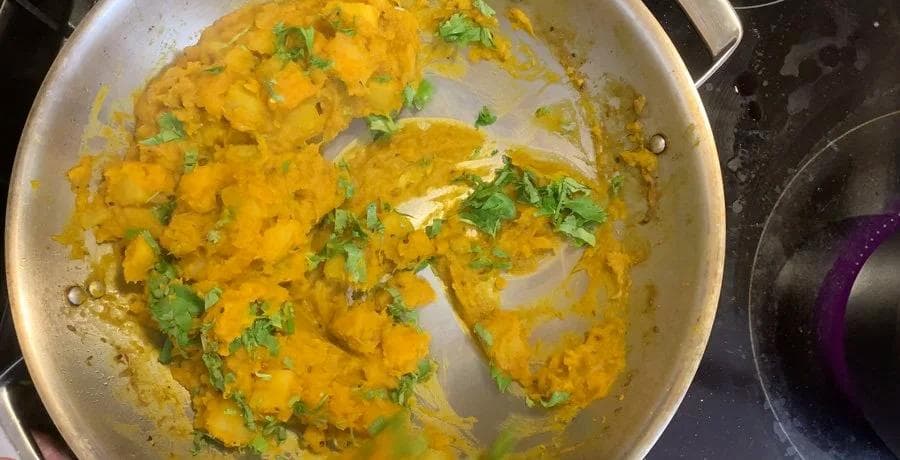Aloo Kohra Sabji curry in a pan topped with fresh cilantro, showing cooked potatoes and pumpkin in golden turmeric sauce.