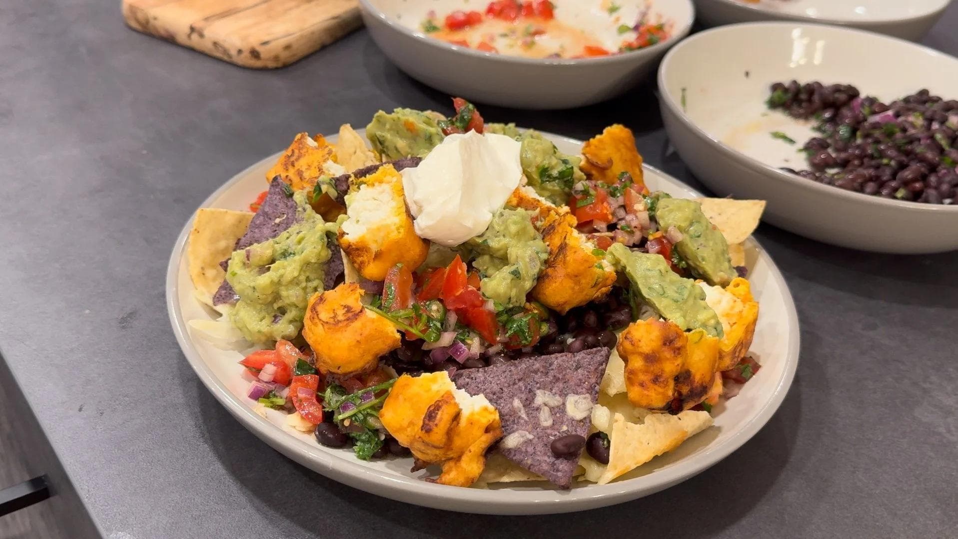 Paneer pakora nachos topped with guacamole, pico de gallo, black beans, sour cream, and fresh cilantro on a white plate.