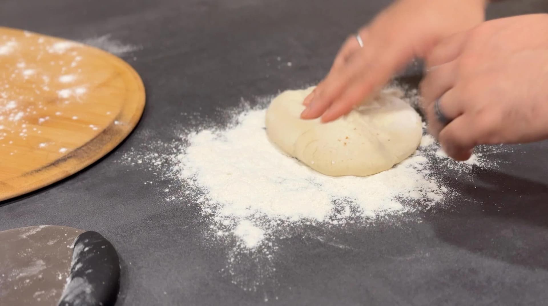 Hands stretching pizza dough on a floured dark countertop next to a wooden board and rolling pin.