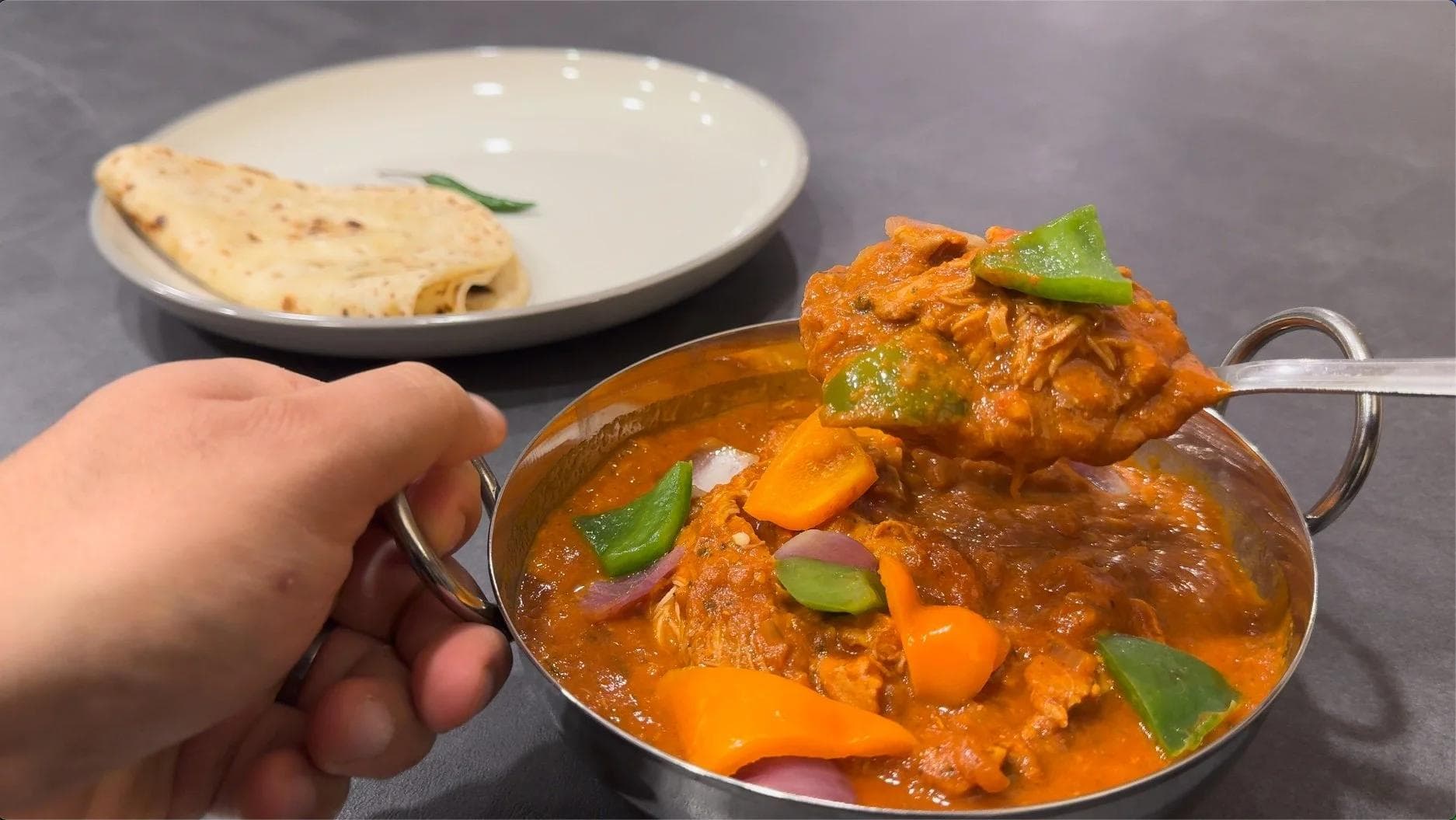 Chicken Tikka Masala being lifted from a stainless steel serving bowl with naan bread on a plate in the background.