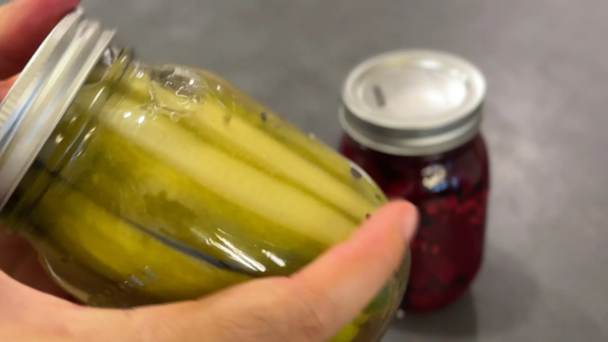 Hand holding a jar of yellow-green pickled vegetables with pickling brine, with a jar of red pickled vegetables blurred in background.