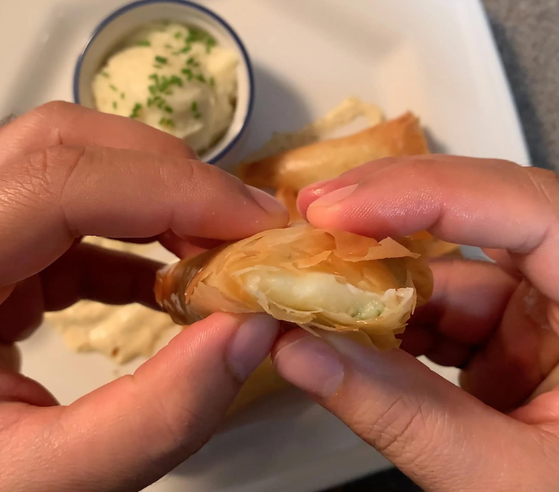 Hands holding a crispy golden-brown samosa with cilantro-mint chutney in a bowl nearby.