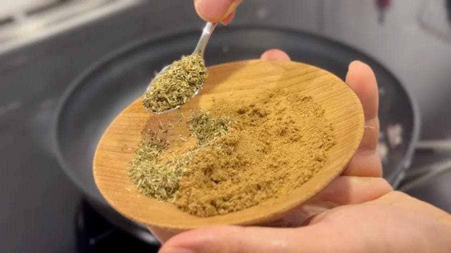 Spoon of cumin powder being added to wooden spoon over pan with sautéed onions and garlic.