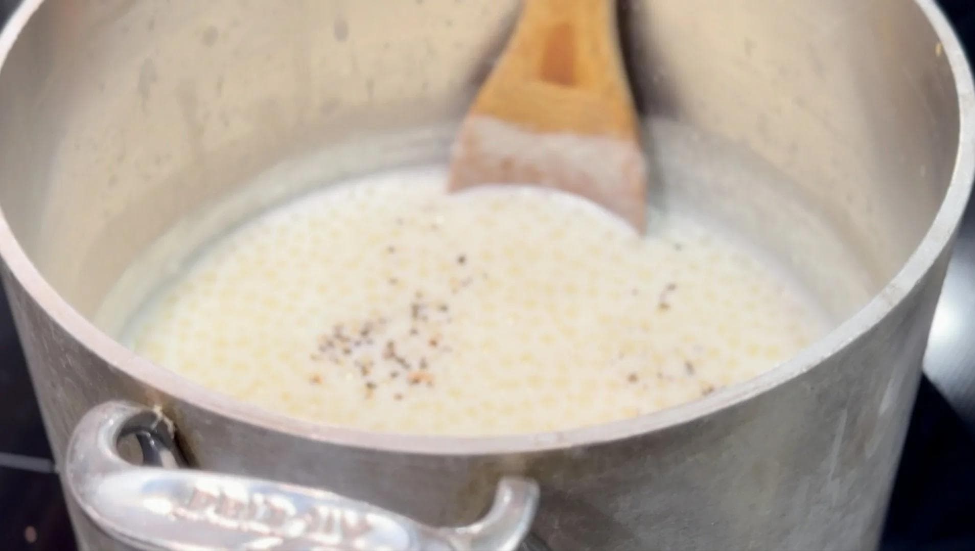Creamy sabudana kheer simmering in a pot with cardamom seeds sprinkled on top, being stirred with a wooden spoon.