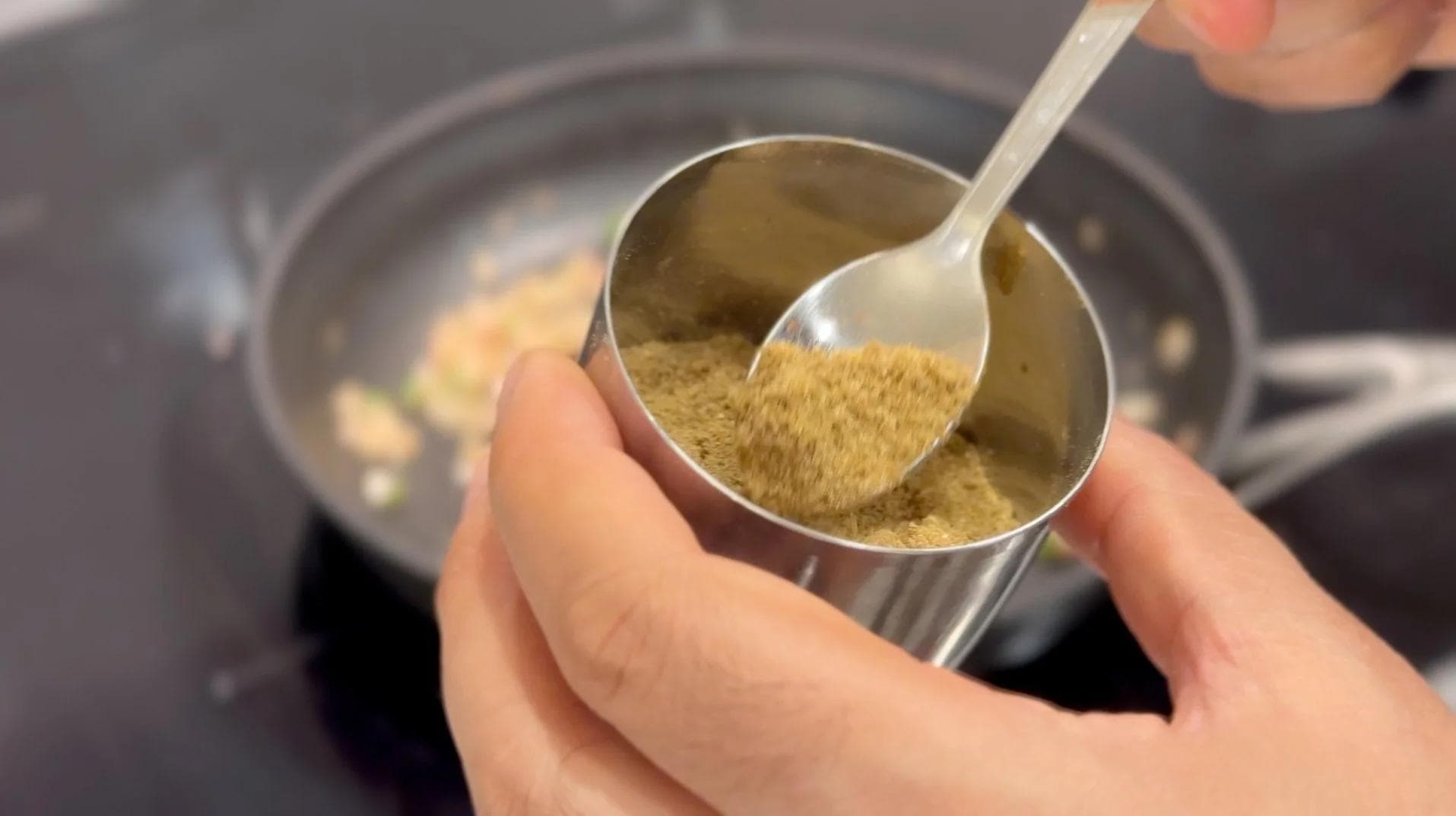 Cumin seeds being measured in a metal spoon over a pan during the sautéing step of mushroom patty preparation.