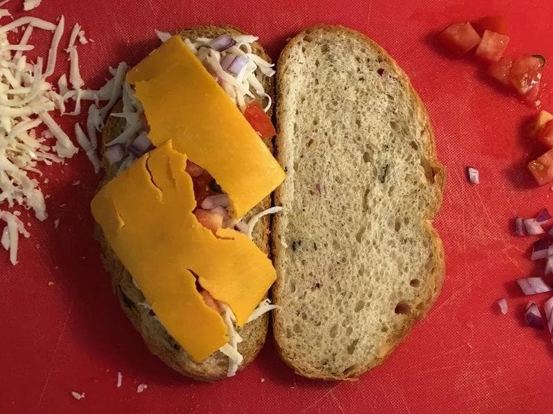 Grilled cheese sandwich with yellow cheddar cheese, shredded mozzarella, diced onions, and tomatoes between wheat bread slices.