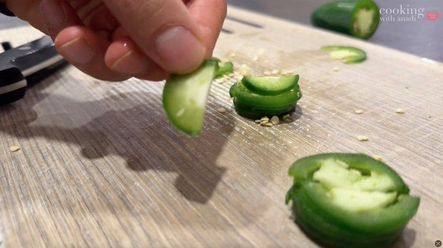 Hand slicing green jalapeño peppers on a wooden cutting board for sushi preparation.