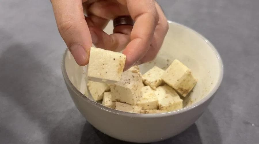 Hand picking up a seasoned tofu cube from a bowl of cubed tofu pieces.