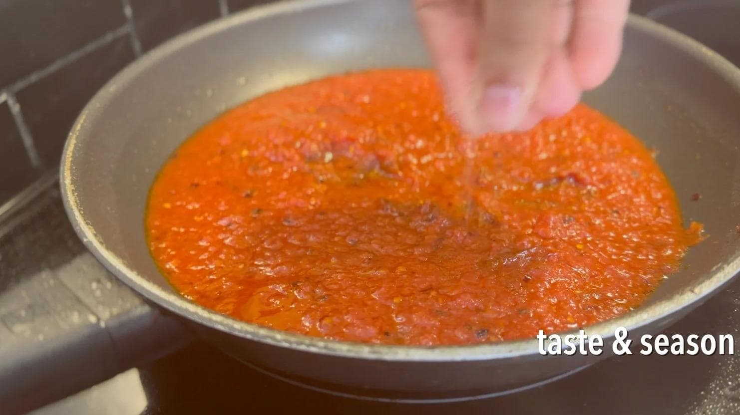 Hand sprinkling seasoning into a pan of bubbling red tomato sauce with oil