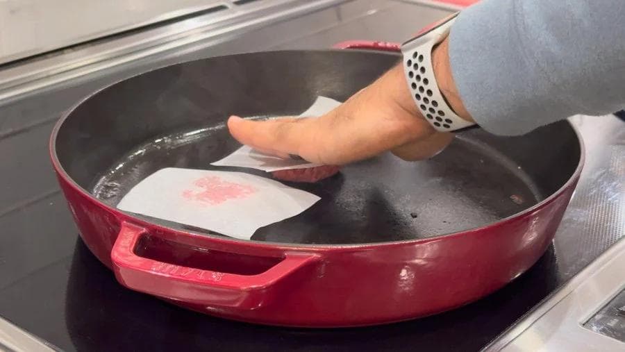 Hand using scraper to flip smash burger patties in red pan on stovetop, parchment paper visible.