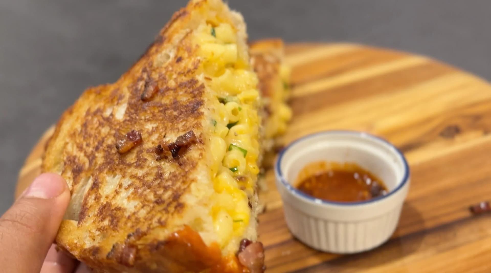 Hand holding a grilled mac and cheese sandwich with melted cheese visible, served with dipping sauce on a wooden board.