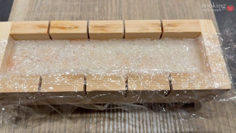 Wooden oshizushi mold with plastic wrap covering pressed sushi, ready for cutting with guide marks visible.