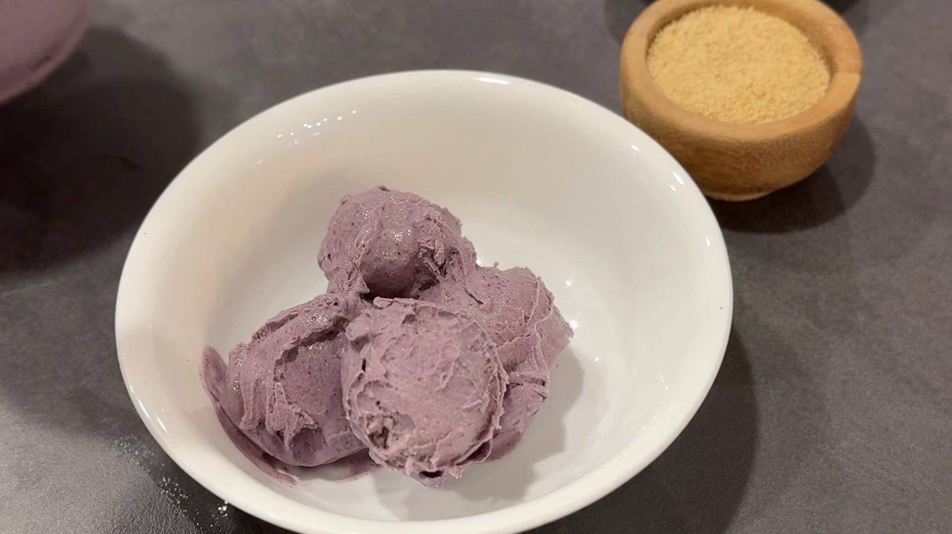 Purple blueberry cheesecake ice cream scoops in a white bowl with graham cracker crumbs in a wooden bowl nearby.
