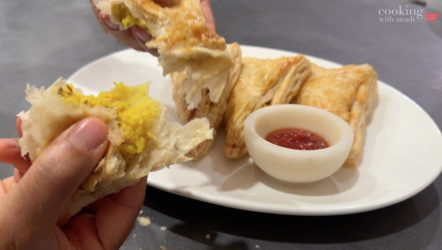 Hand holding a golden, flaky aloo puff patty over a white plate with additional patties and red sauce.