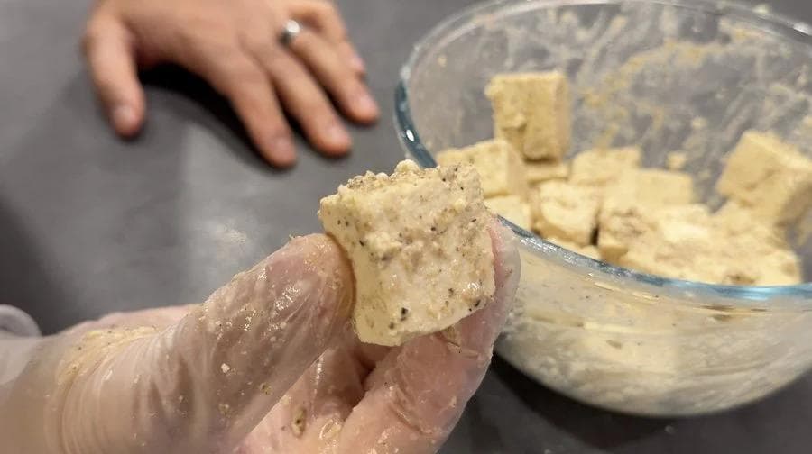 Hand holding a coated tofu cube with a bowl of breaded tofu pieces in background.