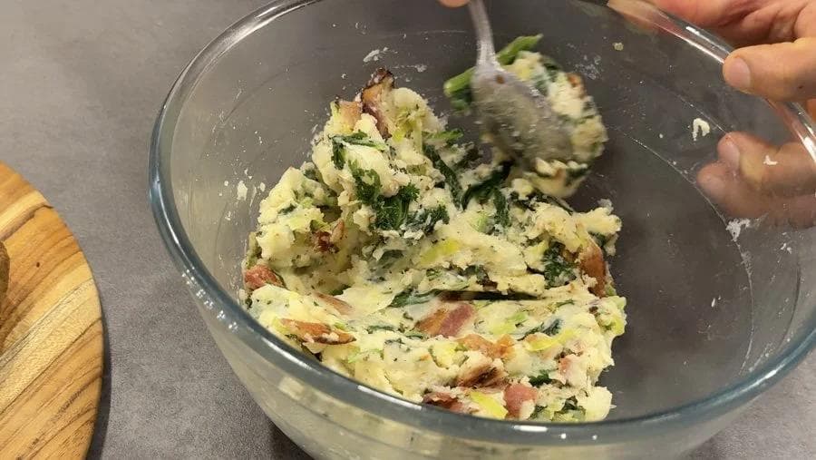 Colcannon filling being spooned into a glass bowl, showing mashed potatoes mixed with bacon and chopped greens.