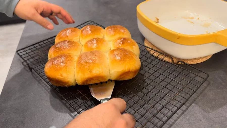 Golden brown dinner rolls on a cooling rack with butter being applied to the top