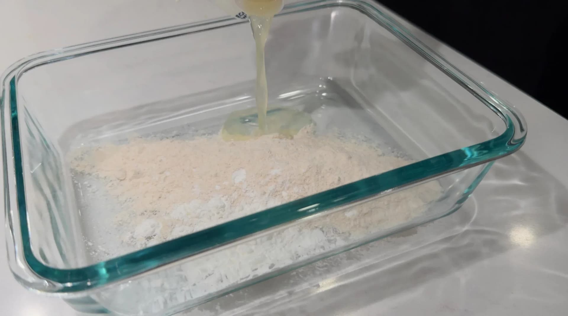 Egg white being poured over a layer of cornstarch and rice flour mixture in a glass baking dish.