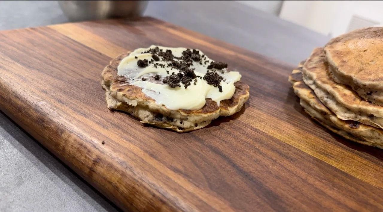 Gluten-free pancakes stacked with cream cheese glaze and topped with crushed Oreos on a wooden board.