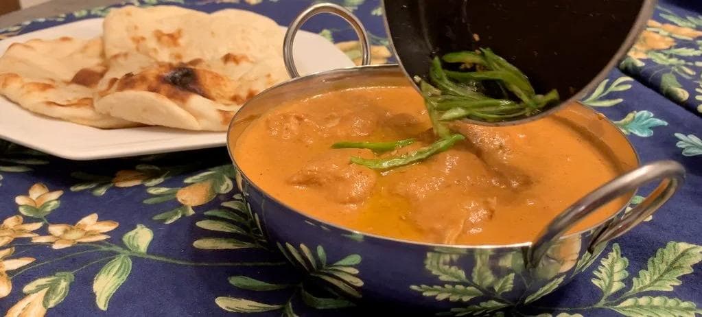 Chicken Changezi served in a decorative bowl with creamy tomato sauce, garnished with fried green chillies and fenugreek leaves, with naan bread on the side.