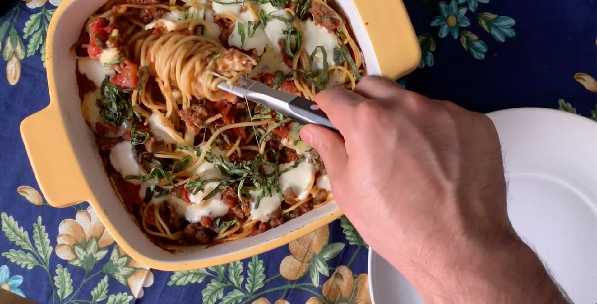 A fork lifting a serving of baked spaghetti casserole with melted cheese and fresh basil from a yellow baking dish.