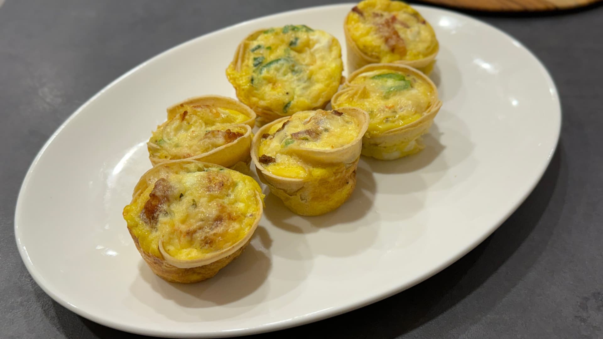 Six baked egg cups with cheese and herbs on a white oval plate against a gray surface.