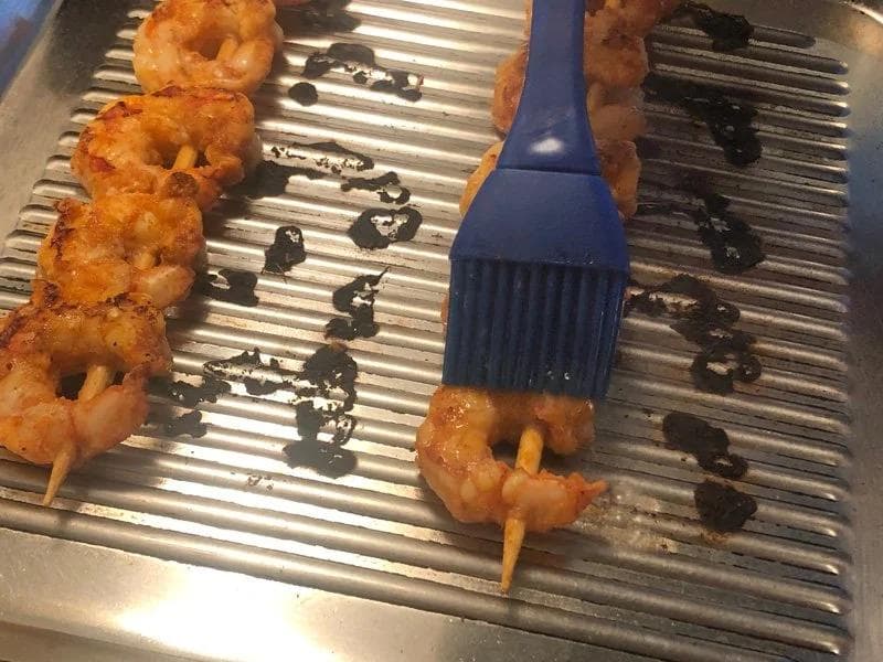 Brushing dark sauce onto grilled shrimp skewers on a ridged grill pan using a blue silicone basting brush.